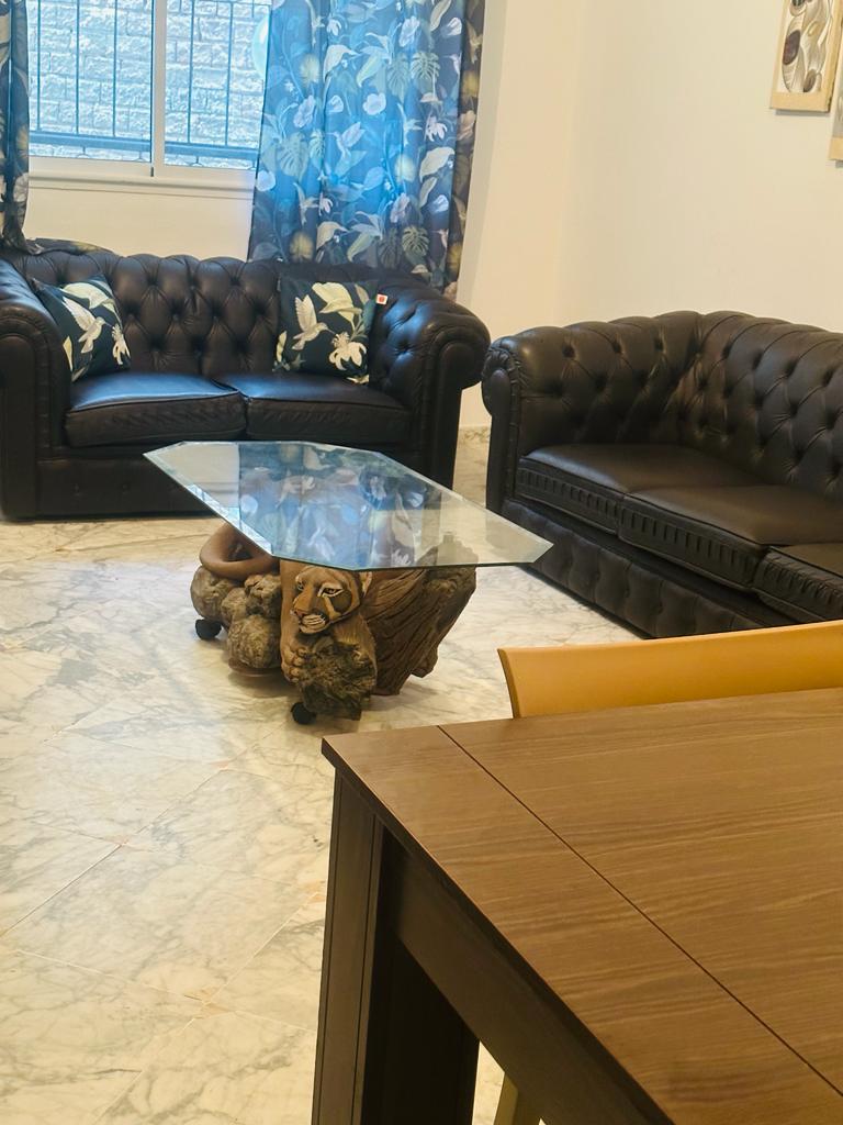 A living area includes two dark leather sofas facing each other, complemented by a unique glass coffee table supported by an animal sculpture. Natural light filters through a window adorned with floral curtains, illuminating the marble flooring and wooden dining table nearby.