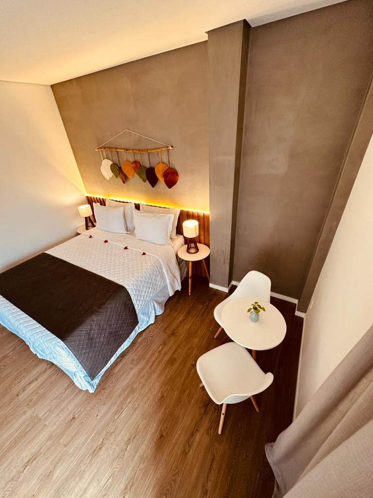 A neatly arranged sleeping area features a spacious bed with a light blanket. A small table and two chairs are positioned beside the bed. Soft lighting is provided by two bedside lamps, and a decorative piece hangs on the wall above the bed.