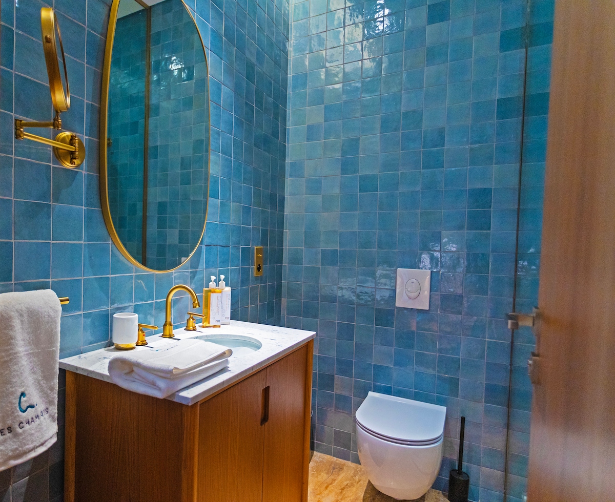 A bathroom is presented with glossy blue tiles adorning the walls, reflecting light. A wooden vanity features a white marble countertop and a round mirror. A modern toilet is positioned adjacent to the vanity, providing a clean and contemporary aesthetic.