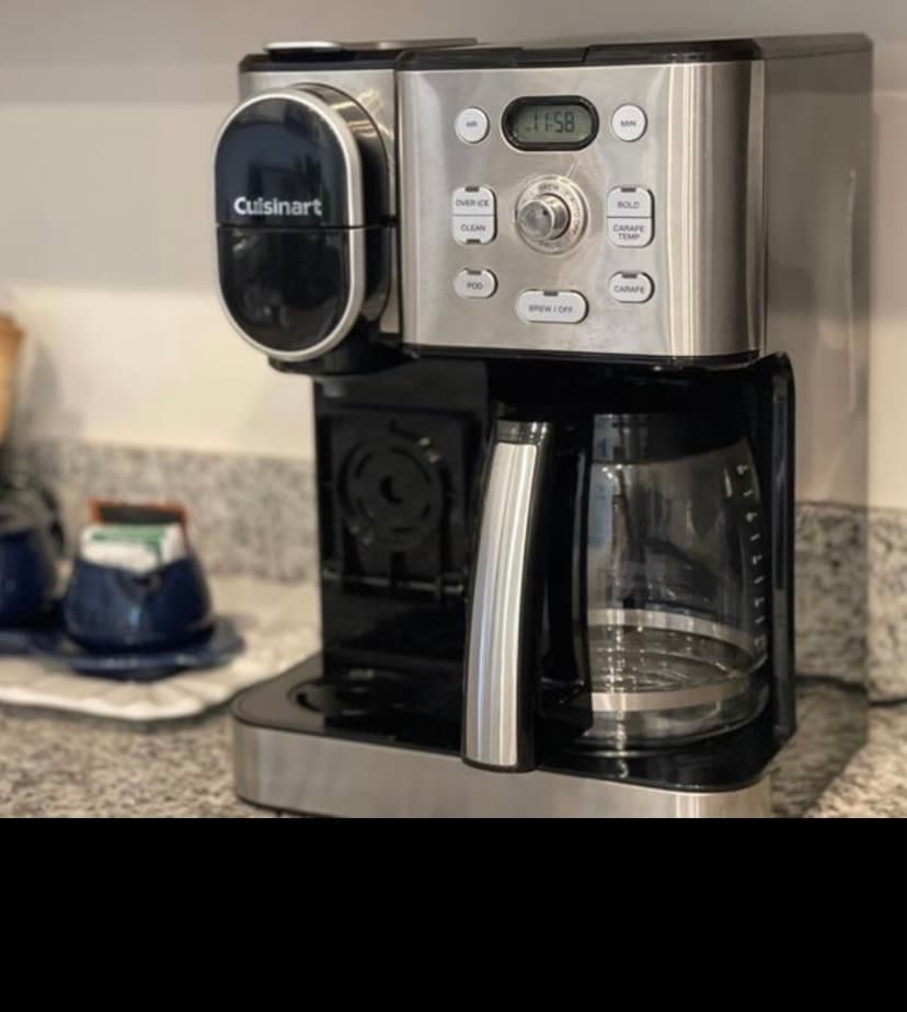 A modern coffee maker sits on a granite countertop, featuring a glass carafe and a digital display panel. The coffee maker is complemented by a small tray holding blue ceramic mugs in the background, adding functionality to the kitchen space.