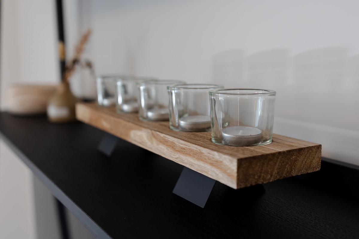 A wooden shelf displays a simple arrangement of five glass tea light holders, each positioned evenly on a rustic wooden base. The background is minimal, allowing the natural textures of the wood and glass to stand out.