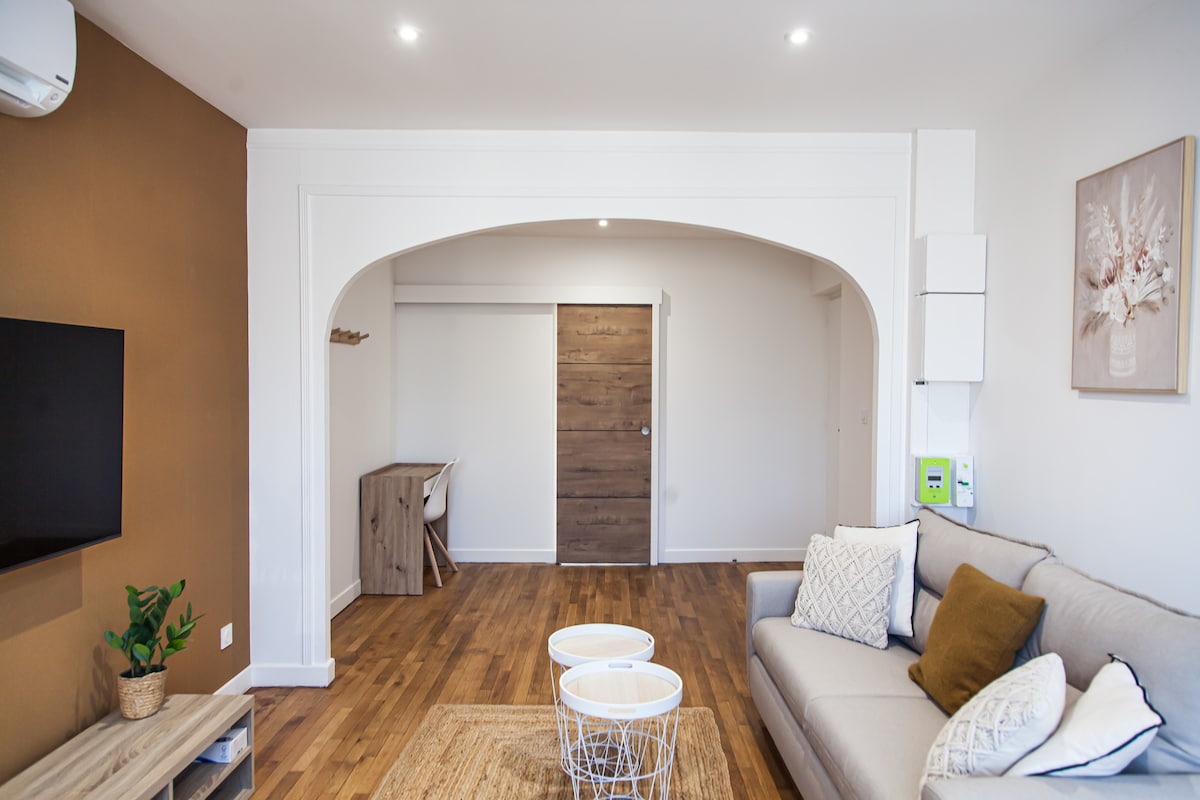 A spacious living area is showcased, featuring a light-colored sofa with decorative pillows. A soft area rug is located beneath a pair of round coffee tables. A wall-mounted TV is visible, and a wooden desk is positioned near the doorway, enhancing the inviting atmosphere.