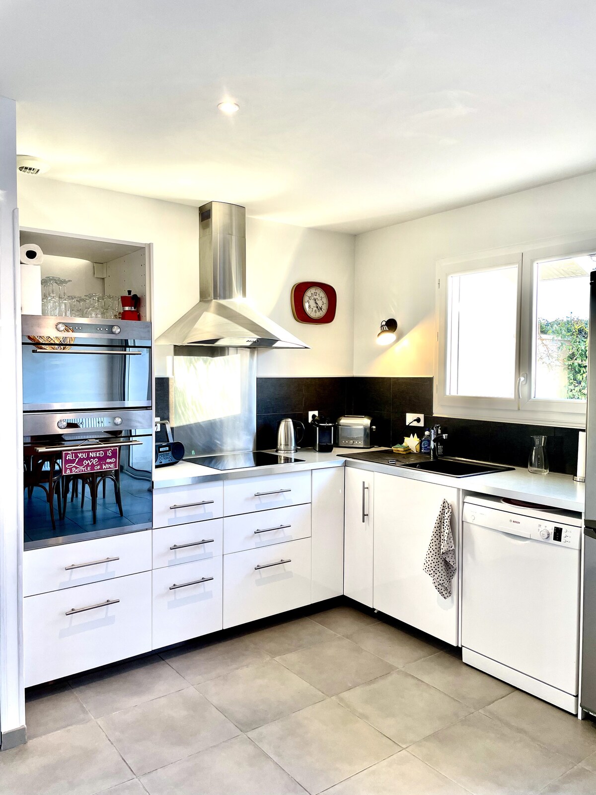 A modern kitchen features sleek white cabinets and stainless steel appliances. A chimney hood sits above the stovetop, while an oven and microwave are built into the cabinetry. Natural light floods in through the window, creating a bright and functional cooking space.