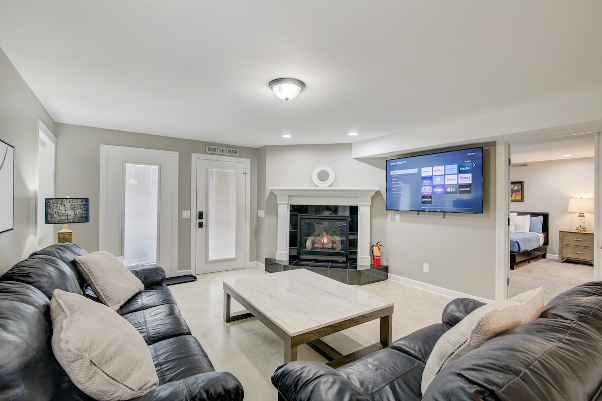A spacious family room features comfortable black leather sofas and a marble coffee table. A wall-mounted Roku TV displays various streaming options. Natural light filters through glass doors leading outside, and a gas fireplace is situated against a neutral-colored wall, enhancing the cozy atmosphere.