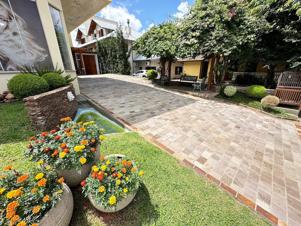 A well-maintained outdoor area features a paved driveway, surrounded by vibrant flower beds in large planters. Lush greenery and trees provide shade, while a small water feature adds tranquility to the inviting space.