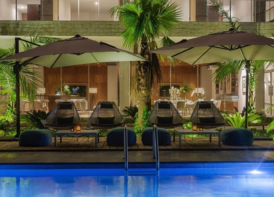 A serene outdoor space is highlighted with loungers beneath large umbrellas beside a swimming pool. Lush green plants and palm trees add to the atmosphere, with a modern backdrop featuring indoor seating and dining areas visible through glass doors.