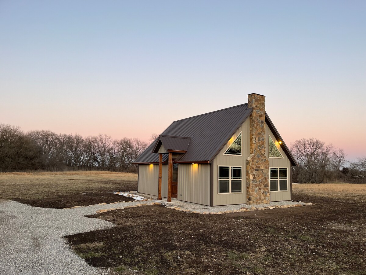 A cabin with a distinctive A-frame design is surrounded by open fields, featuring large windows on either side of a central stone chimney. The exterior blends natural stone and metal siding, while a gravel path leads to the front entrance, illuminated by soft outdoor lighting.