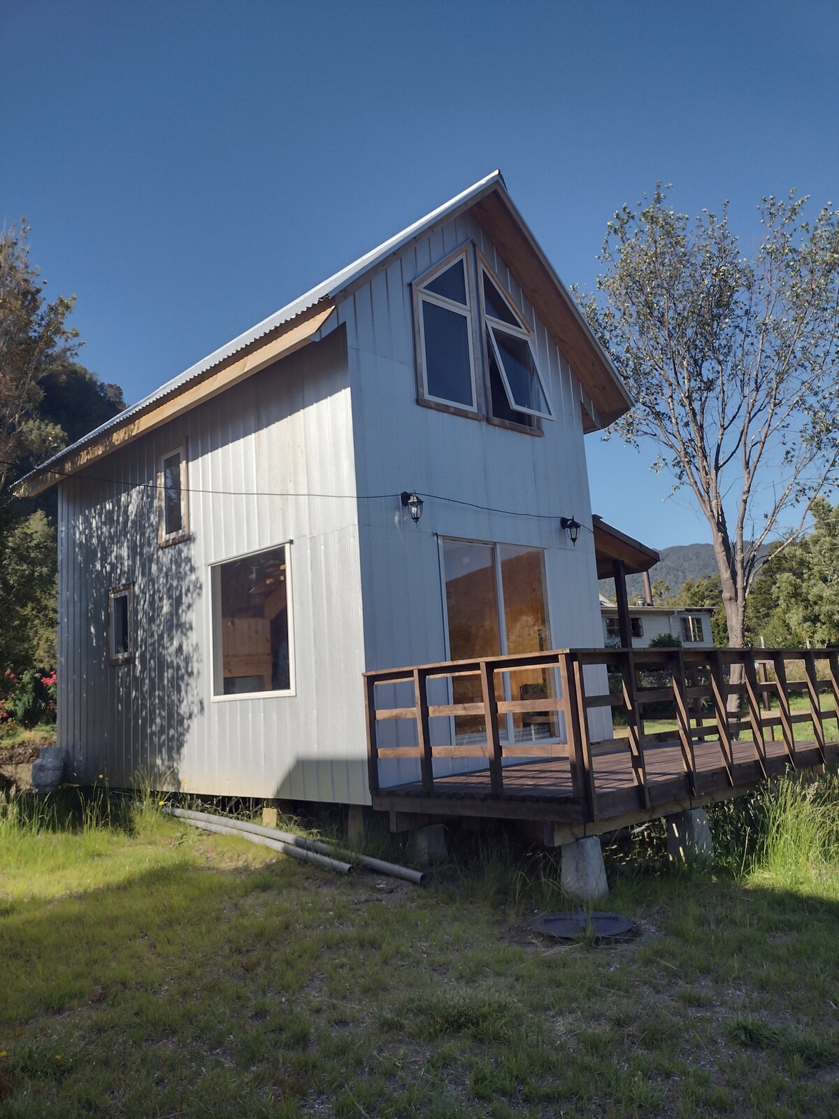 A two-story cabin is shown, featuring a light-colored exterior with large windows that allow natural light. A wooden deck extends from the front, surrounded by lush greenery, providing a peaceful outdoor space. Trees frame the cabin, enhancing the natural setting.