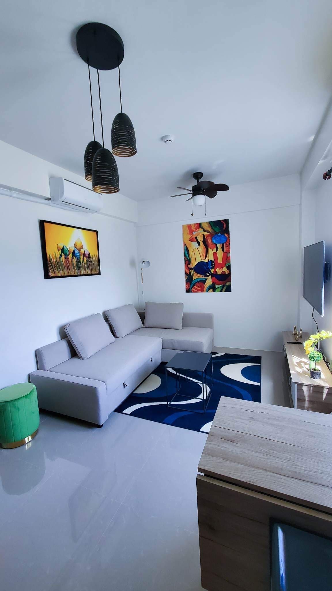A modern living area features a light gray sofa placed against an accent wall. Colorful artwork adorns the walls, complemented by stylish light fixtures overhead. A flat-screen television is mounted and a small table sits in front of the sofa, enhancing the space's functionality.
