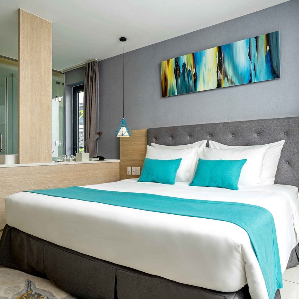 A well-furnished bedroom features a spacious bed with teal accent pillows, complemented by a stylish bed runner. A modern painting hangs above the headboard, while the en-suite bathroom area is partially visible. Soft natural light enters through the window, enhancing the serene atmosphere.