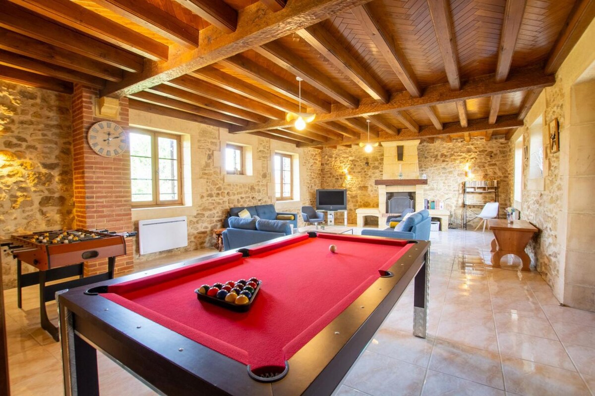 A spacious lounge area features a billiard table and foosball table, with comfortable seating arranged around a modern fireplace. Large windows provide natural light while exposed wooden beams enhance the rustic charm of the stone walls.