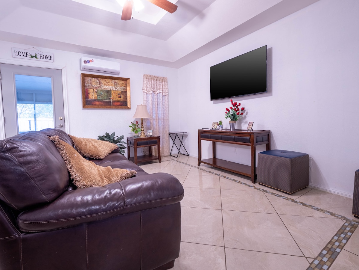 A cozy living space features a brown leather sofa adorned with a plush throw pillow. The television is mounted on the wall above a wooden console table, which is decorated with a few picture frames and a vase of flowers. A ceiling fan adds to the comfort of the room.