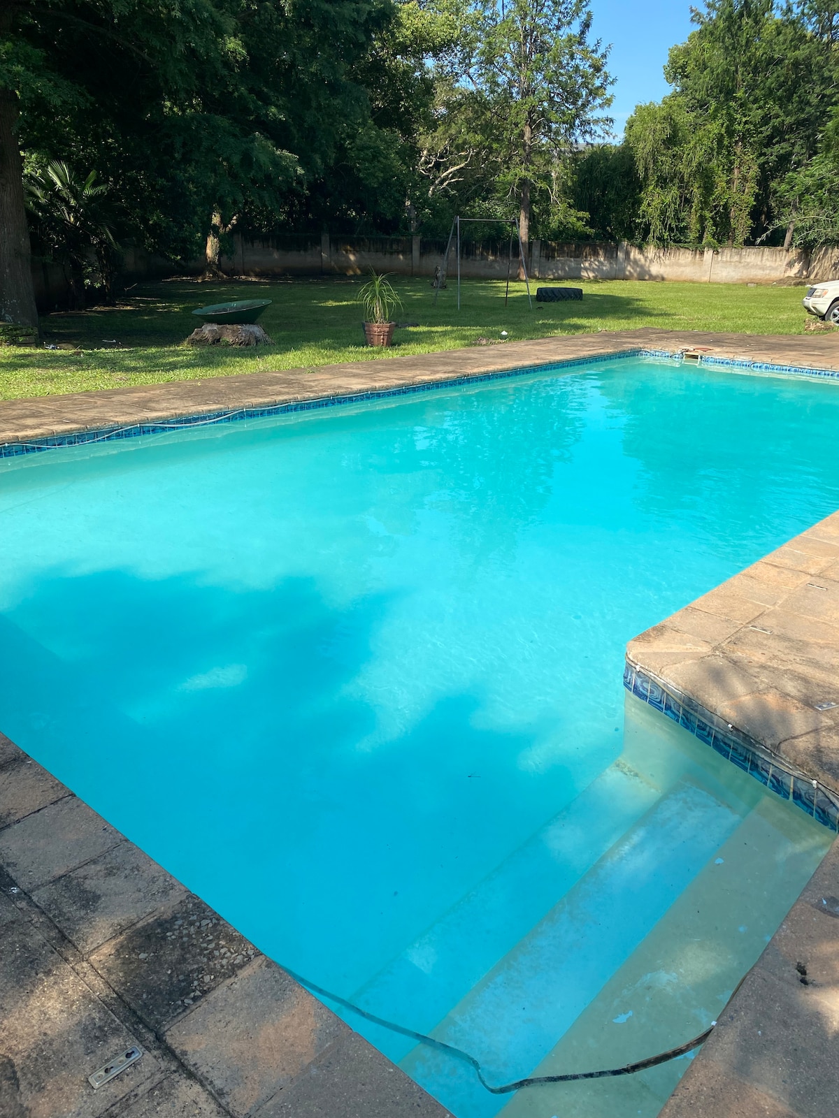 A refreshing swimming pool is nestled in a verdant outdoor space, featuring clear blue water under sunlight. Surrounding the pool, a well-kept lawn and trees create a serene environment, inviting relaxation.