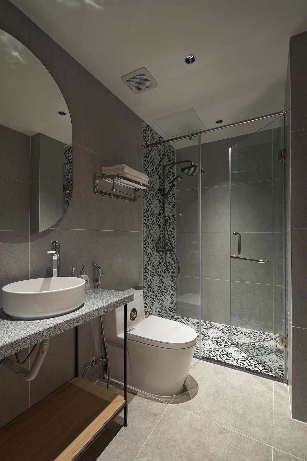 A modern bathroom features a sleek countertop with a circular sink and minimalist faucet. A glass shower enclosure showcases intricate patterned tiles, while a round mirror reflects the stylish design. A wooden shelf holds neatly stacked towels, adding to the organized appearance.