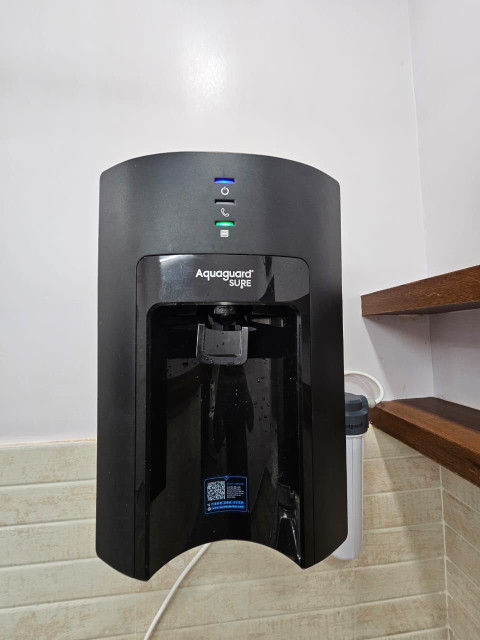 A black water purifier is mounted on the wall, featuring a sleek design with a user interface that includes buttons for different functions. It is positioned above a wooden shelf in a minimalistic kitchen setting.