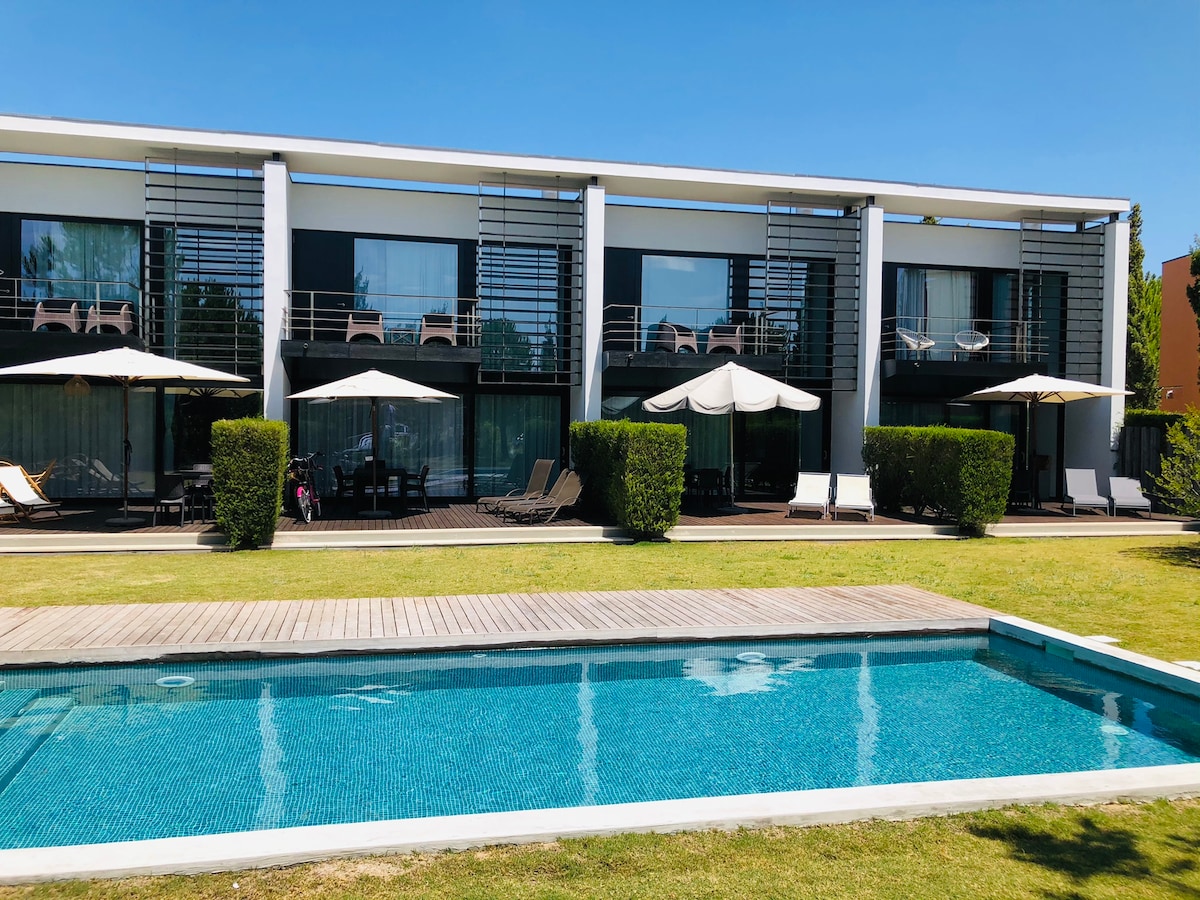 A modern two-story building with large windows overlooks a clear swimming pool. Sun loungers and umbrellas are arranged around the pool area, complemented by manicured greenery and wooden decking. The space invites relaxation in a serene landscape.
