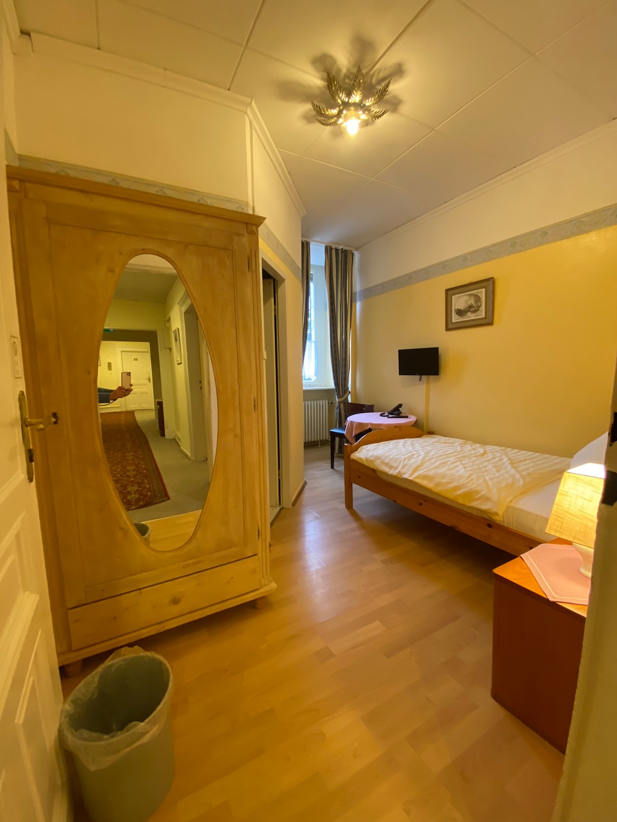 A narrow corridor leads to a cozy bedroom, featuring a spacious bed beside a lamp and bedside table. A large mirrored wardrobe reflects the warm wooden tones, while a small wall-mounted TV is positioned at the foot of the bed. Natural light enters from a window at the far end.