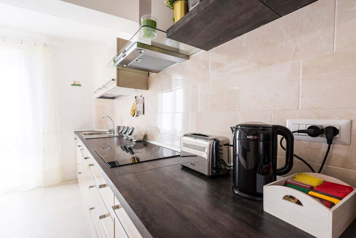 A modern kitchen is presented with sleek cabinetry and a spacious countertop. Appliances include a kettle, a toaster, and an induction cooktop. Natural light filters through a nearby window, enhancing the clean and functional design. A small container holds colorful kitchen items, adding a touch of vibrancy.