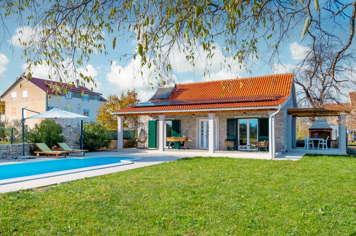 A modern villa with a red-tiled roof is surrounded by lush greenery. The spacious yard features a swimming pool and a shaded area with seating. Large glass doors offer views of the enclosed outdoor space, while nearby houses are visible in the background.
