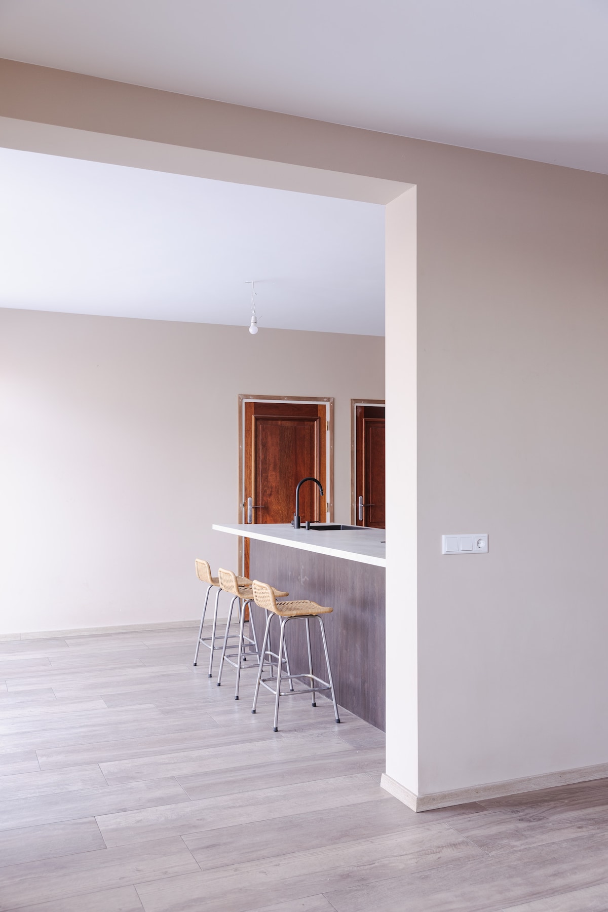 An open kitchen area features a breakfast bar with three wooden stools. The light-toned walls and flooring enhance the spacious feel. Wooden cabinetry is visible in the background, and a modern faucet is positioned above the countertop, providing a functional yet simple design.