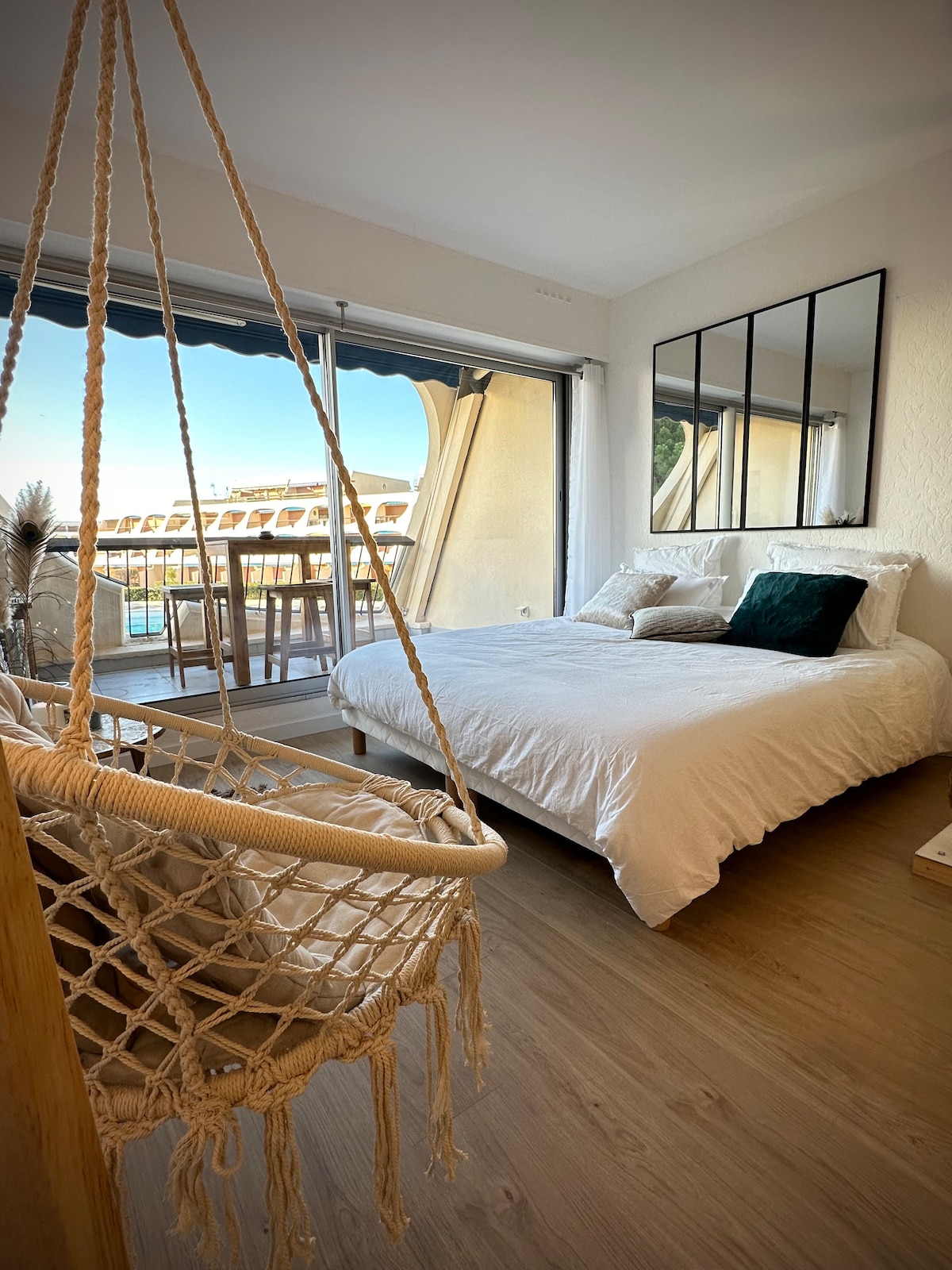 A cozy bedroom features a spacious bed with white bedding and accent pillows. A macramé hanging chair adds a unique touch. Floor-to-ceiling glass doors allow natural light and provide a view of the terrace overlooking the port and sea.