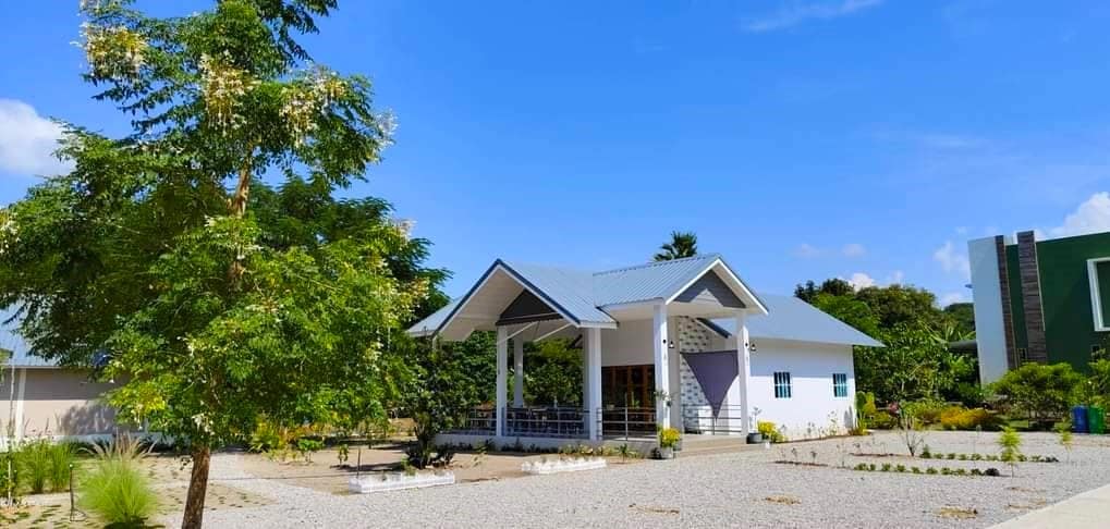 A modern building with a sloped roof and open-design seating area is surrounded by well-maintained landscaping. The space appears bright and airy, benefiting from ample natural light. A large tree provides shade in the foreground, while the surrounding area features gravel pathways and green plants.