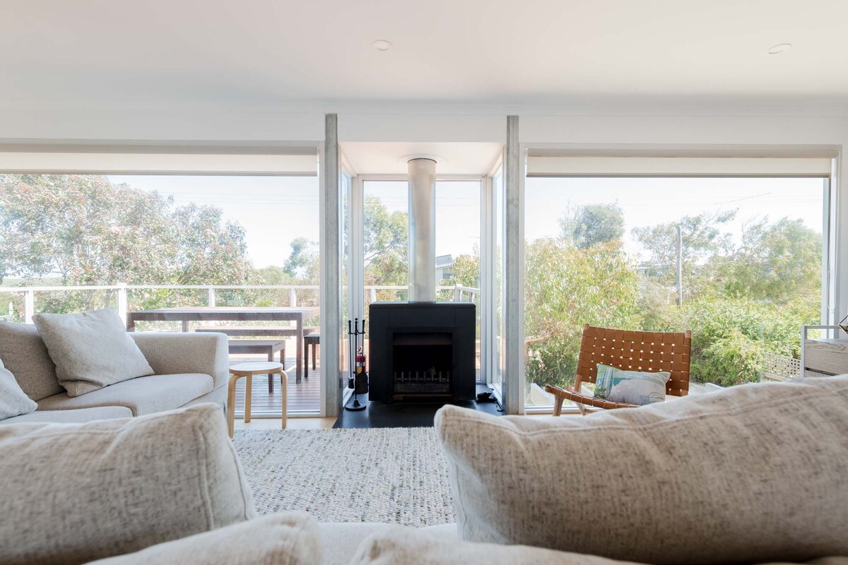 The open-concept living area features a cozy fireplace framed by large glass windows, providing scenic views of the surrounding greenery. Soft, light-colored sofas are placed invitingly, promoting relaxation and comfort in the serene atmosphere.