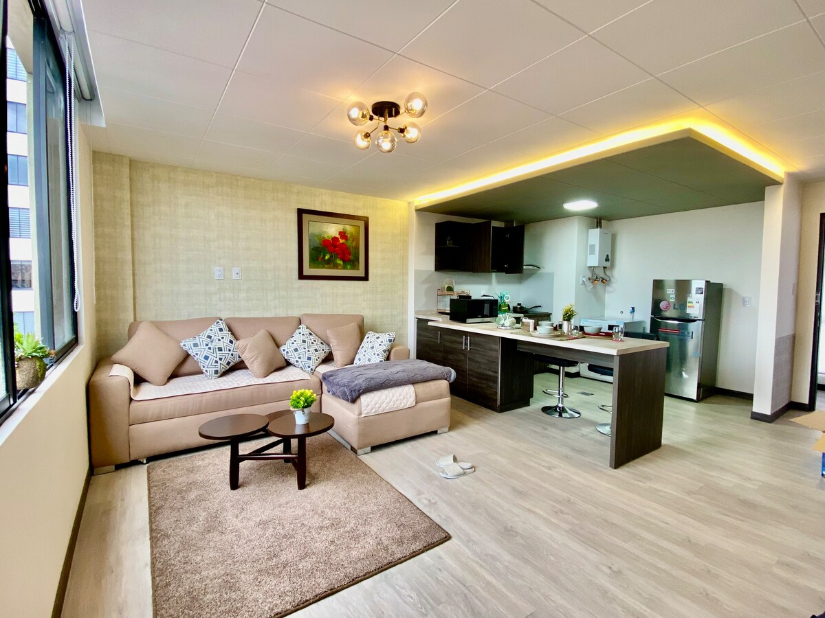 A spacious living area features a comfortable sofa adorned with patterned cushions, paired with a small coffee table. The open kitchen is equipped with modern appliances and a dining space is visible, illuminated by soft overhead lighting and natural light from large windows.