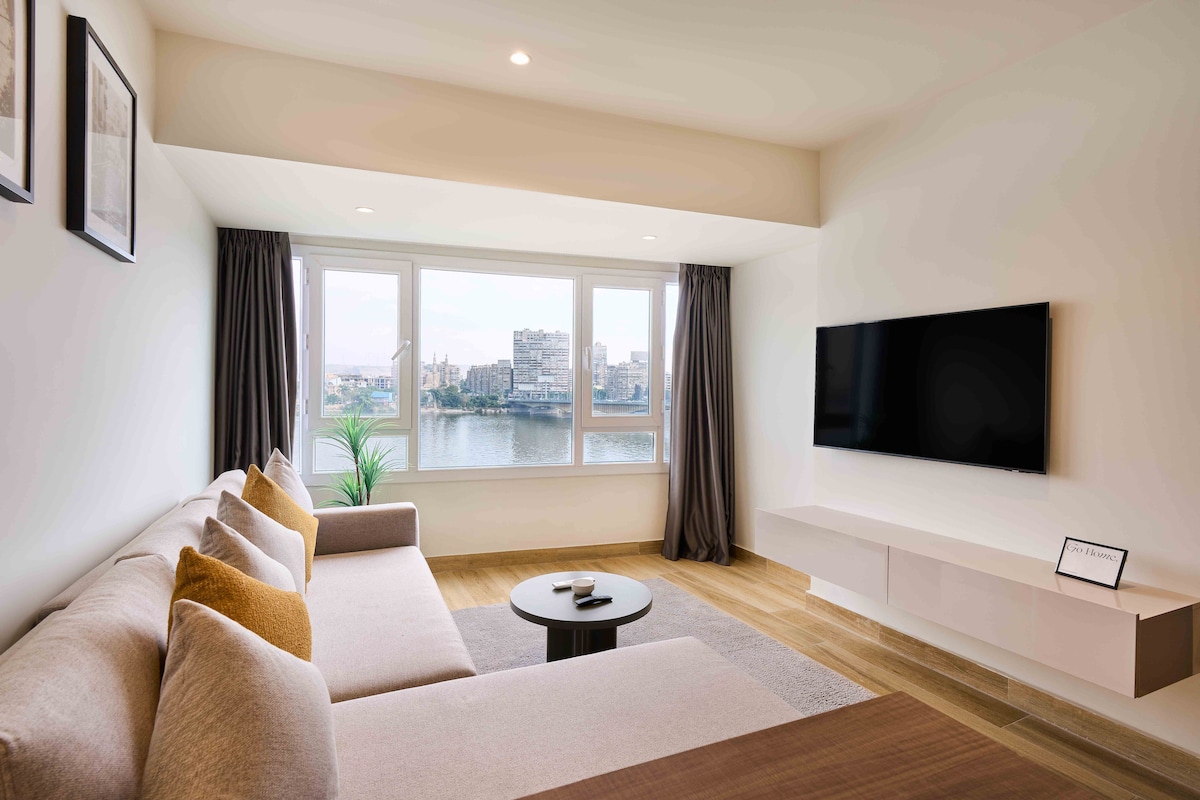A bright living area is showcased with large windows offering views of the Nile River. A comfortable sofa is positioned beside a minimalist coffee table. A wall-mounted television is present, and subtle decor enhances the modern design.