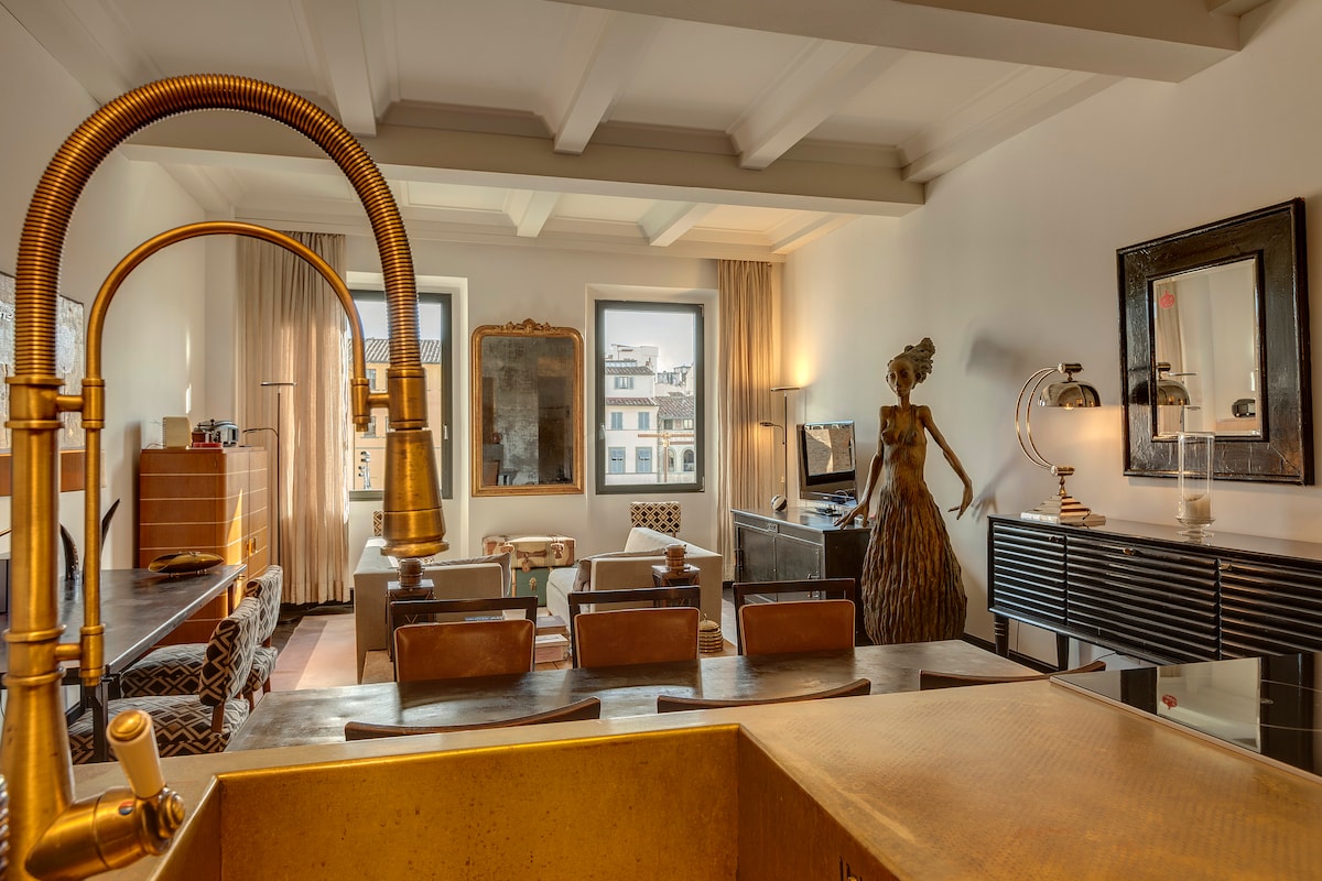 A modern living area features a spacious table surrounded by chairs, with large windows allowing natural light. A decorative statue adds an artistic touch, while stylish furnishings complete the inviting atmosphere. The kitchen counter is visible in the foreground, highlighting a contemporary gold faucet.