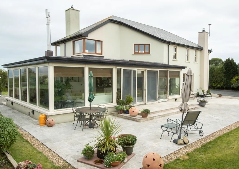 The exterior of the house features a spacious patio area, adorned with potted plants and garden furniture. Large windows allow natural light into the interior, while a green umbrella provides shade over a circular table with chairs. The structure is framed by a well-maintained grassy area.
