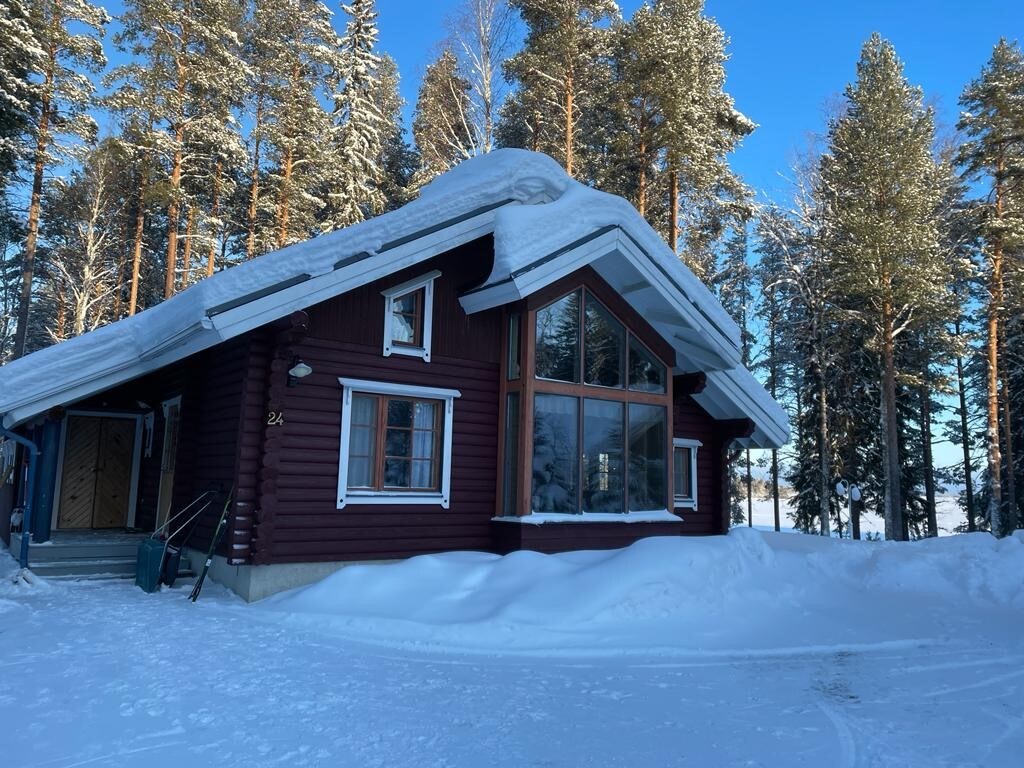 The exterior of a wooden cabin is showcased against a winter landscape, surrounded by snow-covered trees. Large windows provide views of the surroundings, and a gently sloped roof is adorned with snow. A clear path leads to the entrance.