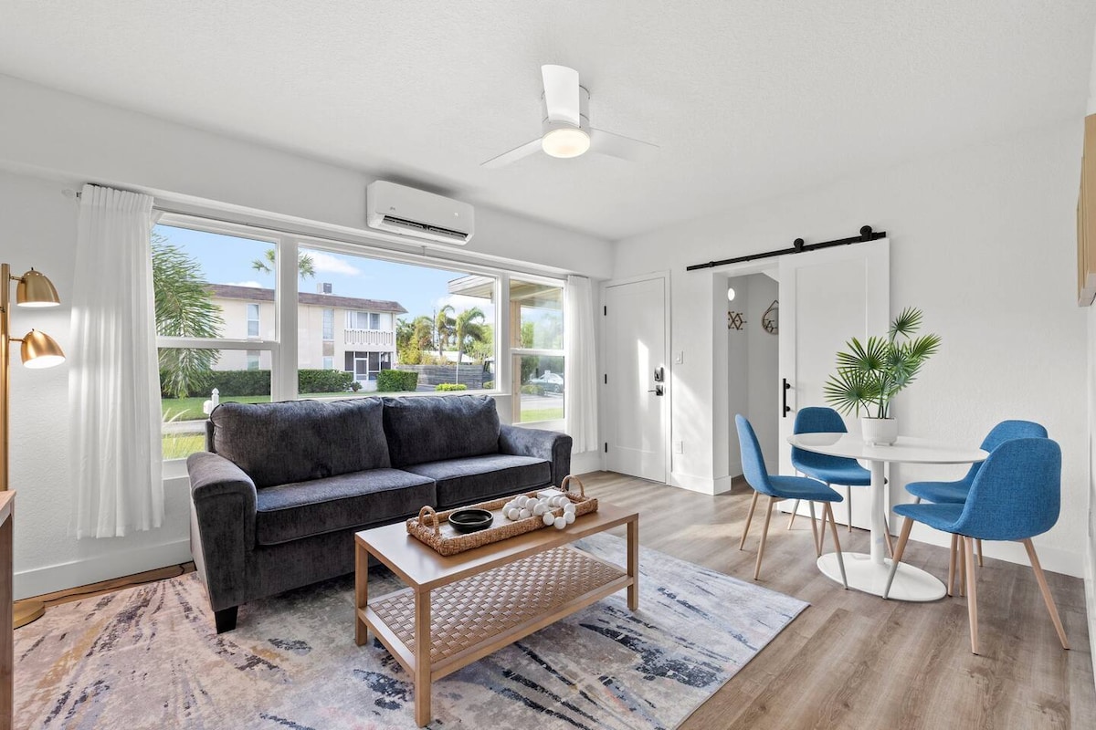 A cozy living area features a dark gray sofa contrasted by light wood accents. A coffee table with decorative items sits centrally. Blue dining chairs surround a small white table, illuminated by natural light from large windows showcasing outdoor greenery.