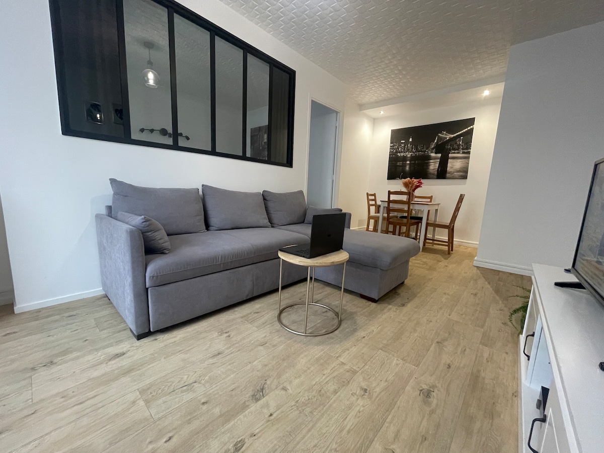 A spacious living area is presented, featuring a light grey sectional sofa, a small round side table, and a laptop resting on it. A dining table for four is visible in the background, accompanied by a stylish wall art piece depicting a cityscape.
