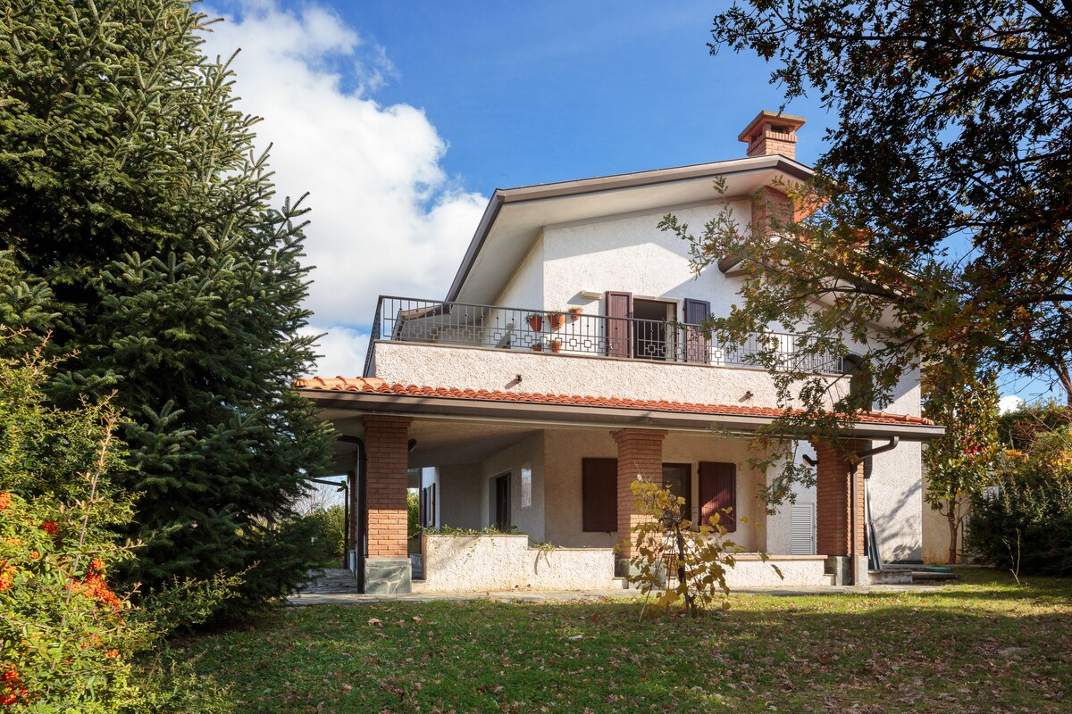 The exterior of a two-story residence is depicted, featuring a covered porch supported by brick columns. A spacious balcony extends from the upper floor, framed by wooden shutters. Lush greenery surrounds the property, contributing to a peaceful outdoor environment.