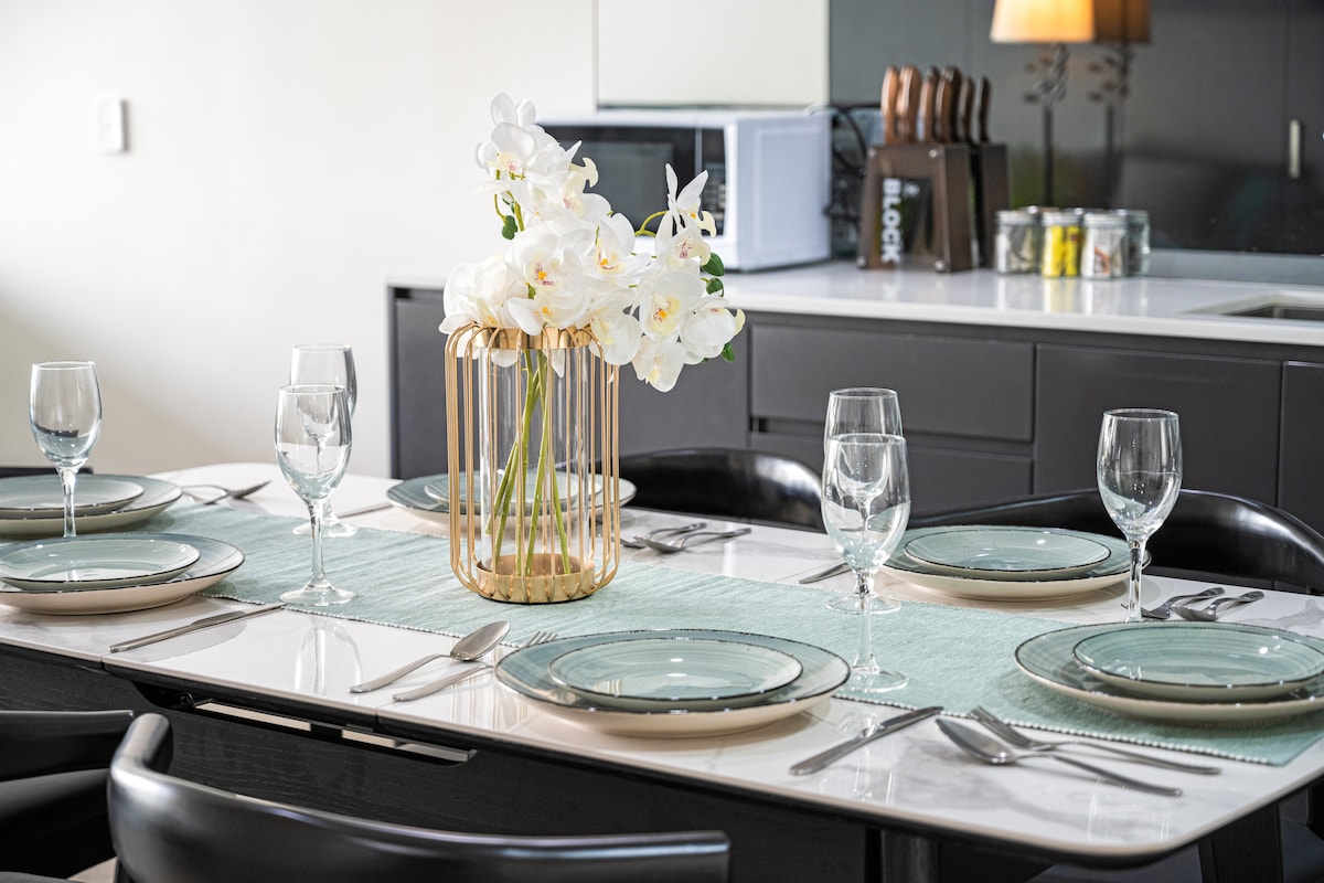 A modern dining table is set for six, featuring elegant glassware and plate settings. A decorative vase holds white orchids at the center, placed on a soft blue table runner. The background includes a sleek kitchen area with stylish cabinetry.