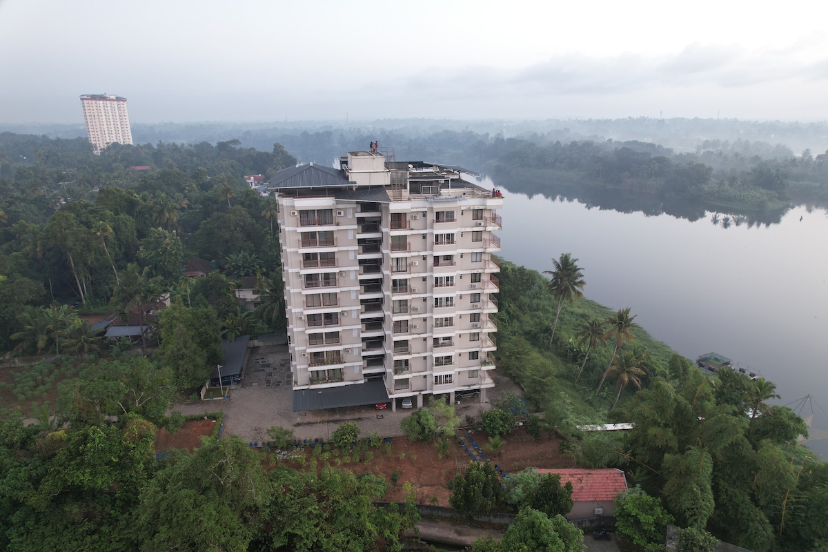 An aerial view showcases a modern multi-story building set against a backdrop of lush greenery and the calm Periyar River. The building's structure is highlighted, and the serene landscape reflects the tranquility of the surrounding nature.