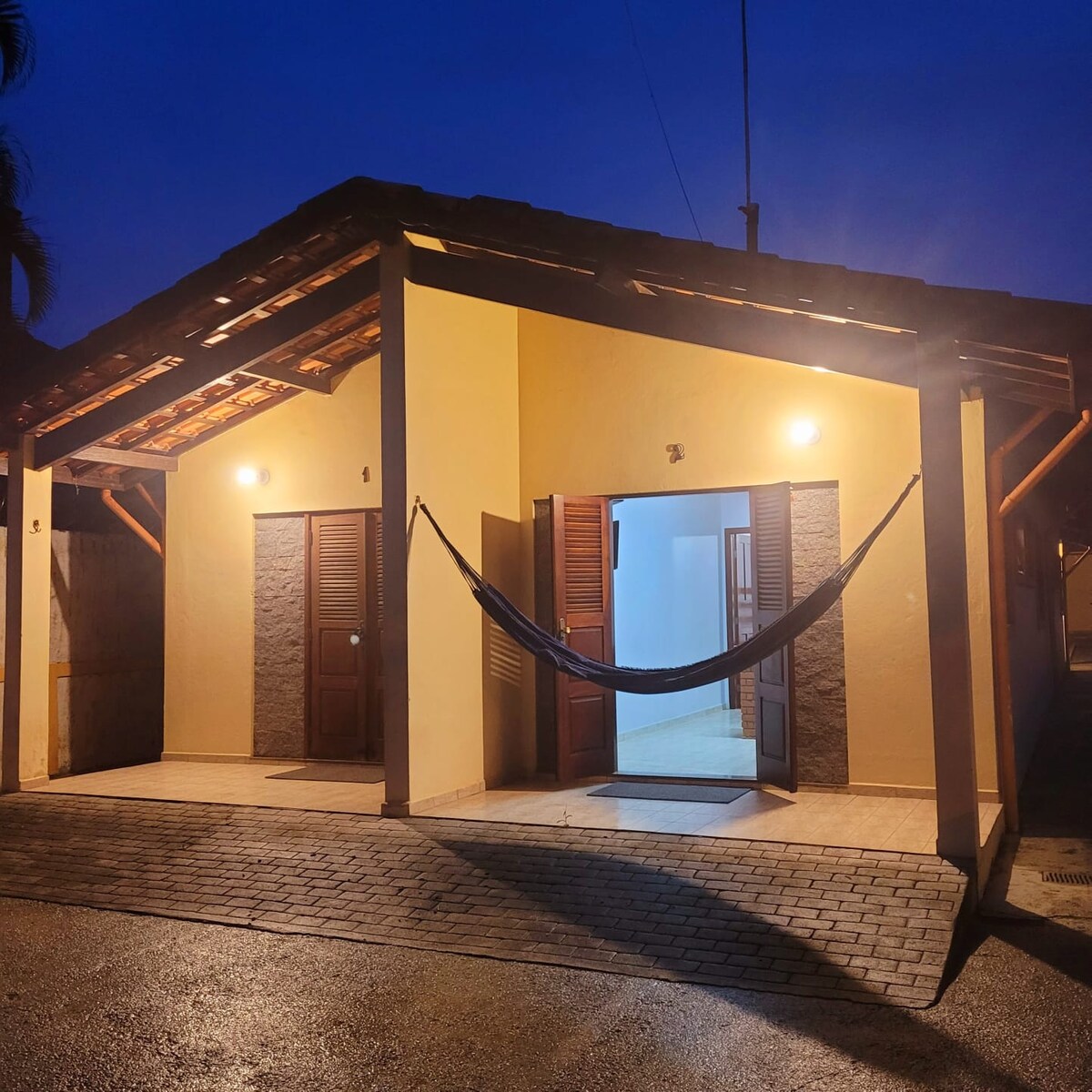 The shared entrance to the chalets is illuminated by soft lighting, featuring a hammock suspended between two support beams. Each door is framed by wooden shutters, and the smooth stone pathway leads to the inviting interiors that are visible beyond the open doors.