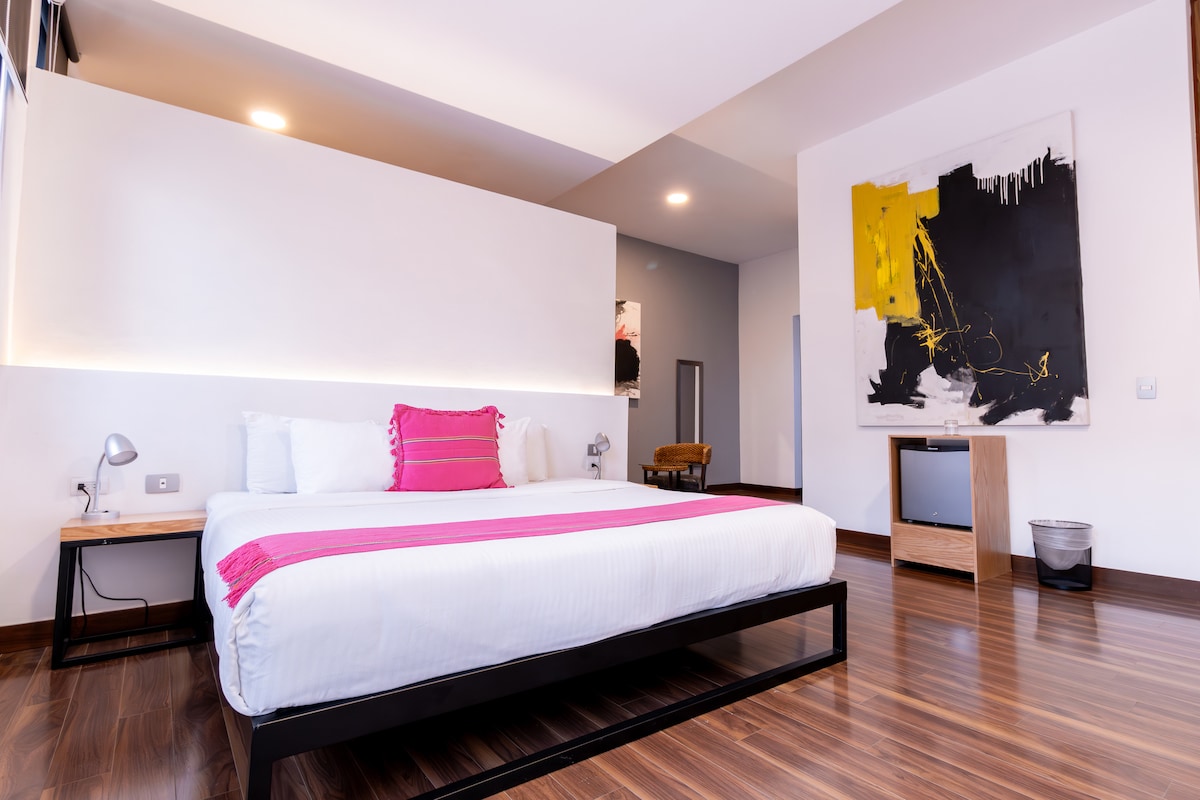 A well-lit bedroom features a spacious bed with white linens and colorful accent pillows. A modern artwork adorns the wall, complemented by a wooden bedside table and a small television. The warm wooden floor adds to the serene atmosphere of the room.