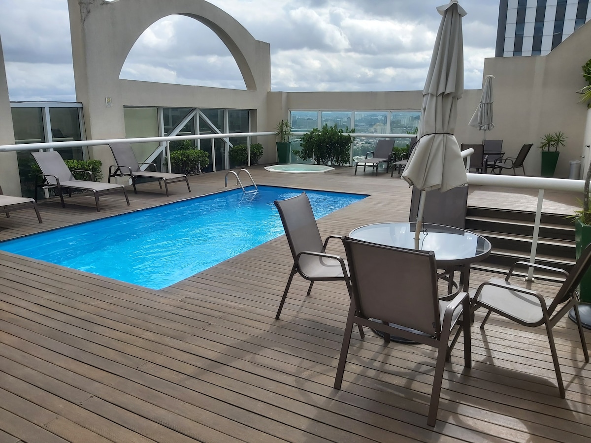 A rooftop pool area is surrounded by wooden decking, featuring a clear blue swimming pool. Several lounge chairs are positioned nearby, along with a small table and umbrella for shade. Potted plants add greenery to the space, enhancing the overall ambiance.
