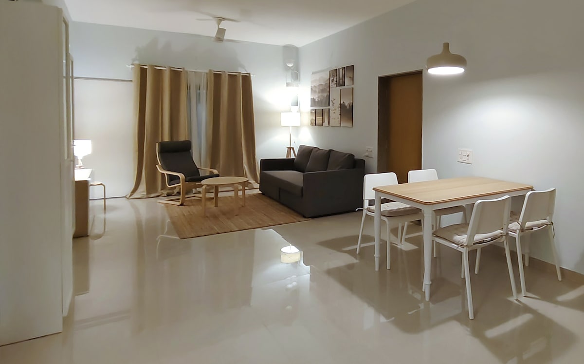 An open living area features a sleek black sofa facing a light wooden table surrounded by white chairs. A natural fiber rug lies beneath the seating arrangement. Soft lighting from a floor lamp and pendant light creates a calm ambiance, while neutral curtains frame the window.
