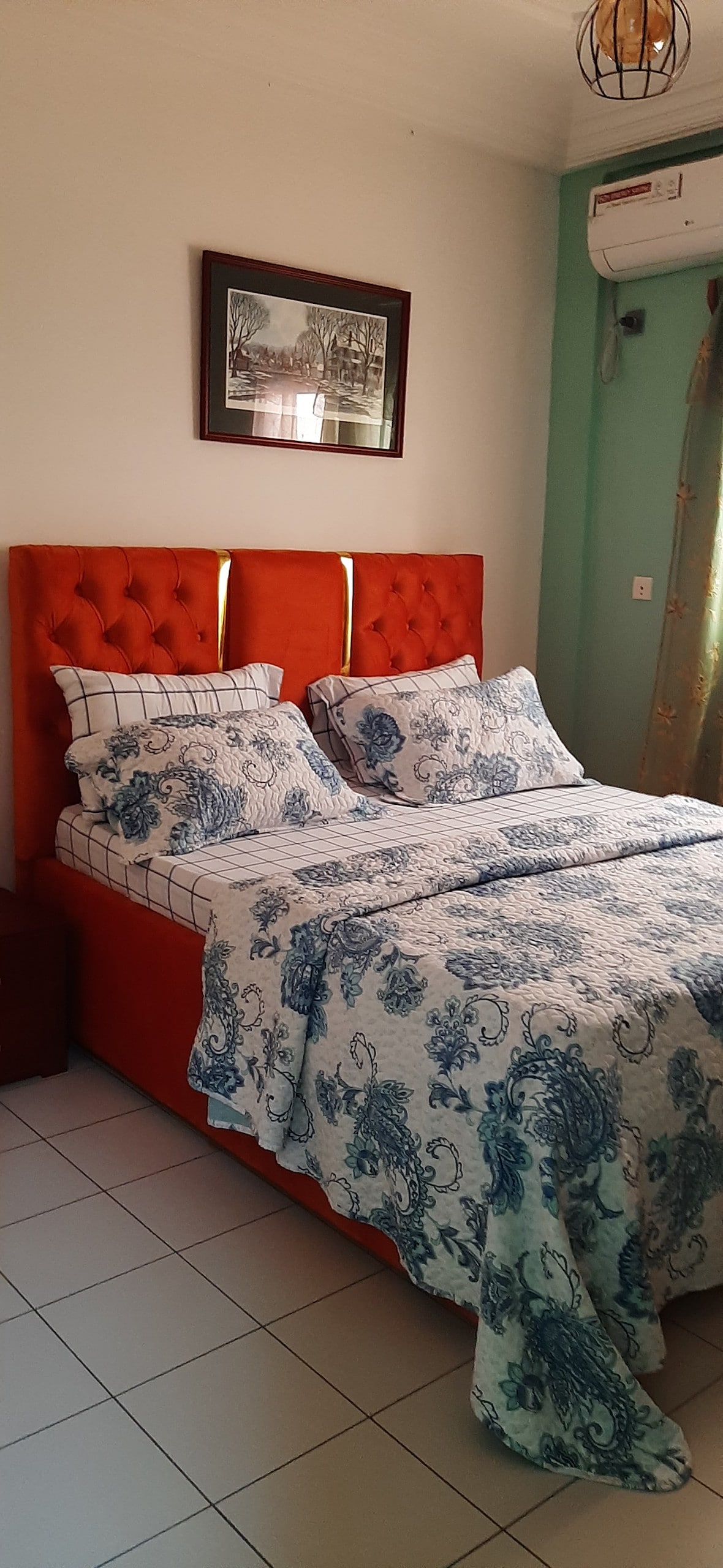A cozy bedroom features a plush orange headboard against a light color wall. A spacious bed is covered with a blue and white patterned comforter, complemented by matching pillows. A framed artwork hangs above the bed, and natural light filters in through a nearby window.