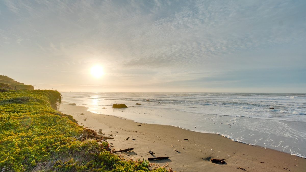 The image captures a serene beach scene at sunset. Soft waves gently lap against the shore, framed by lush greenery. The sun casts a warm glow over the sandy expanse, creating a peaceful atmosphere. The ocean extends toward the horizon, enhancing the natural beauty of the coastline.