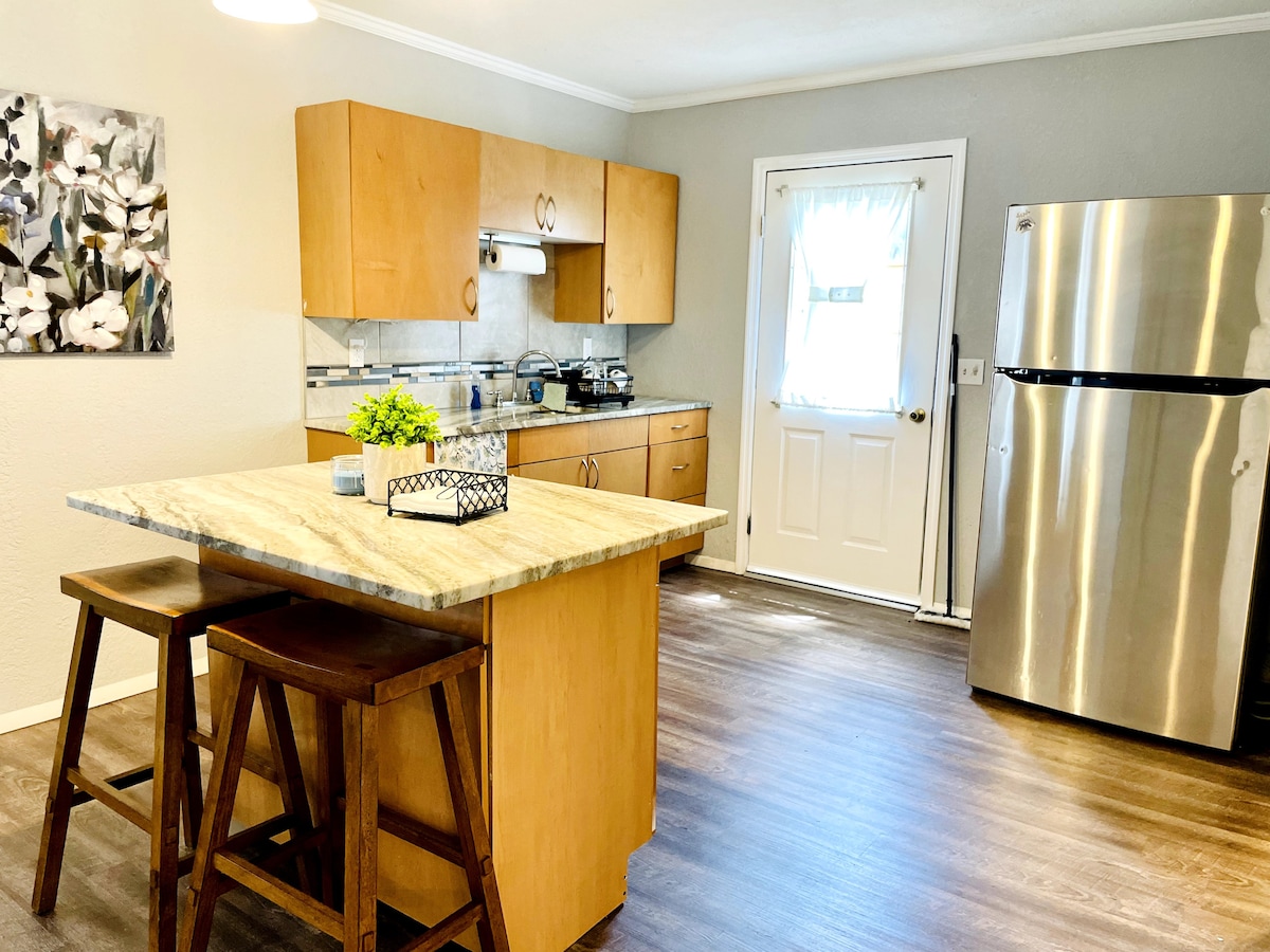 A modern kitchen features light wood cabinetry and a spacious island with a striking granite top. Stainless steel appliances are visible, including a refrigerator, and natural light enters through a door leading outside, complemented by a simple window treatment.