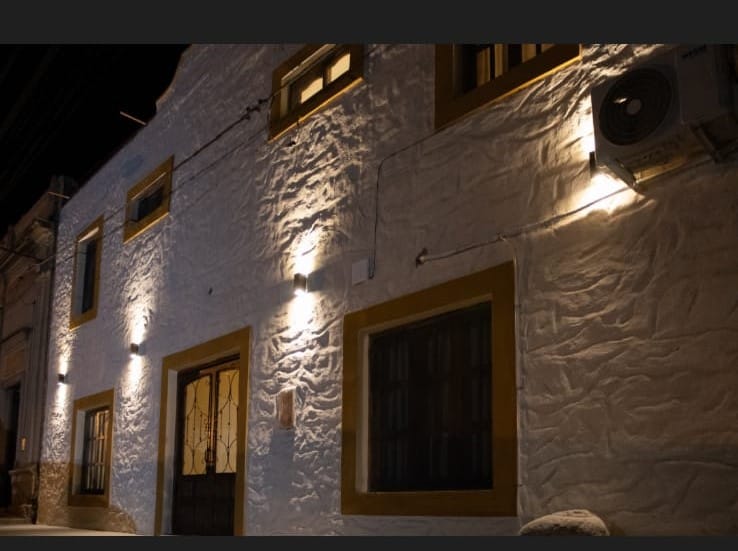 The exterior of the property is showcased with a textured white wall, illuminated by soft lighting. Golden accents around the windows and door add a distinct charm to the facade. Air conditioning units are discreetly positioned on the wall.