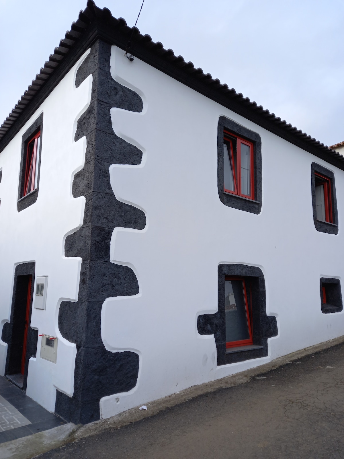 The exterior of the house showcases a combination of white walls and dark stone accents. Bright red window frames contrast with the monochromatic facade, while a simple entrance is visible at the front. A flat roofline provides a clean silhouette against the sky.