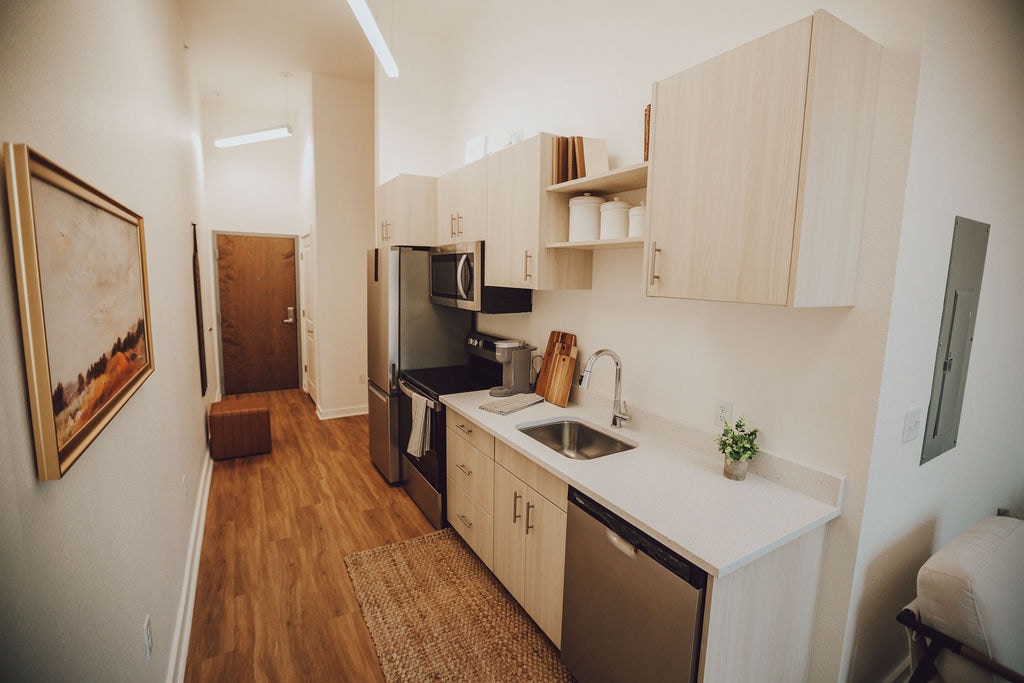 A sleek kitchen area features light-colored wood cabinetry and modern appliances. A stainless steel sink rests between a refrigerator and an oven, with utensils neatly arranged. A small plant adds a touch of greenery, complementing the spacious layout and warm wooden flooring.