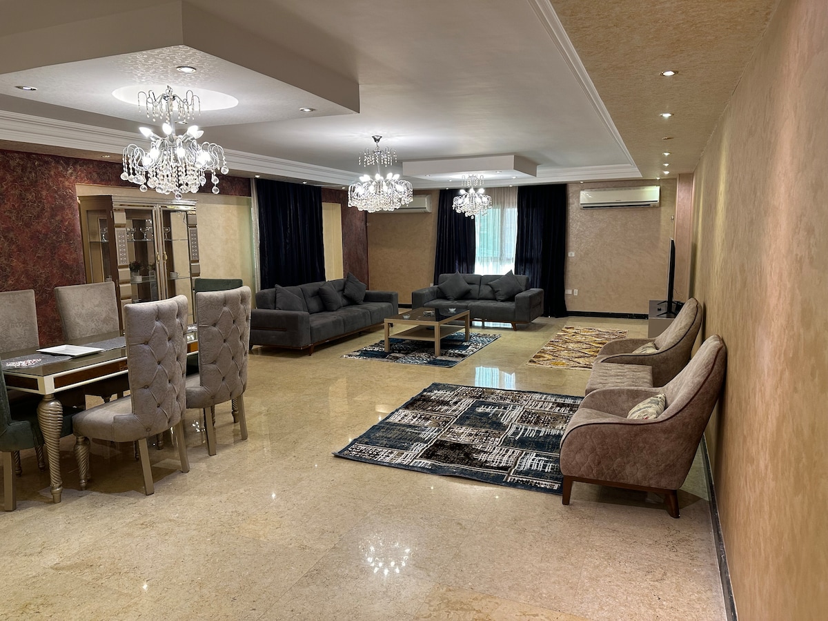 The spacious reception area features elegant furnishings, including a large dining table surrounded by upholstered chairs. Soft sofas are arranged atop patterned area rugs, while multiple chandeliers provide ambient lighting. Dark curtains frame the windows, creating a serene atmosphere.
