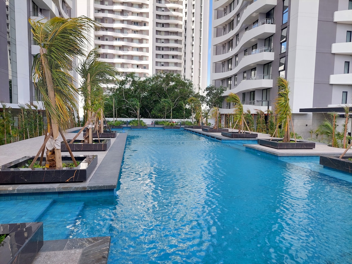 A large outdoor pool is surrounded by tall palm trees and neatly arranged lounge areas. The water reflects the sunlight, creating a serene environment. The modern buildings in the background add a contemporary touch to the tranquil setting.