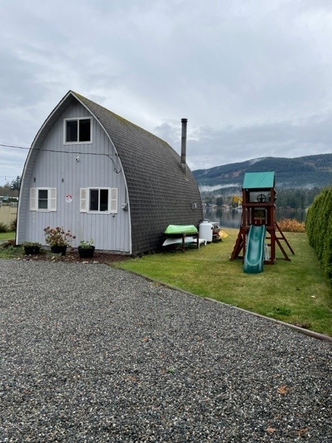 A rustic cabin is shown with a unique arched structure under a cloudy sky. The property features a spacious yard with a playset including a slide, surrounded by neatly trimmed shrubs. Gravel leads up to the cabin, enhancing its quaint charm.